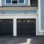 A modern house with light blue siding and white trim features a double garage with dark paneled doors. Two lantern-style lights are mounted above the garage. The driveway is paved in black asphalt. A small shrub is visible to the side.