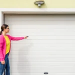 A person wearing a pink cardigan and jeans points a remote at a closed white garage door. The background is a light-colored wall with a small security light above the door.