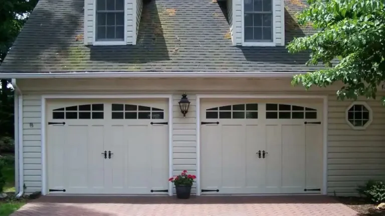 A two-car garage with white doors and black accents is shown. Each door has windows at the top. Above are two dormer windows. A lantern-style light fixture is centered between the doors. A small potted plant with flowers is in front.